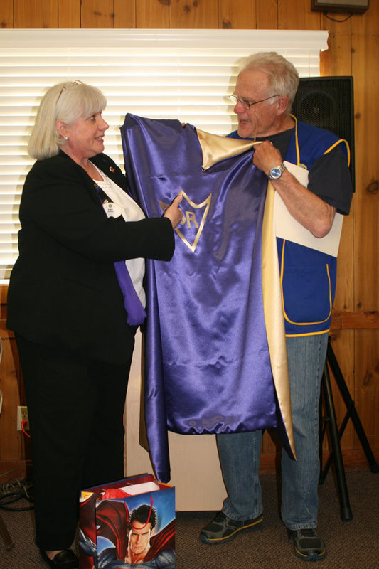 Image: Bonnie Johnson, the District Governor of the Northern Utah Lions club, presents Richard Criddle a super hero cape for his decade of service in the Lions club as chair- man of the eye glass division.